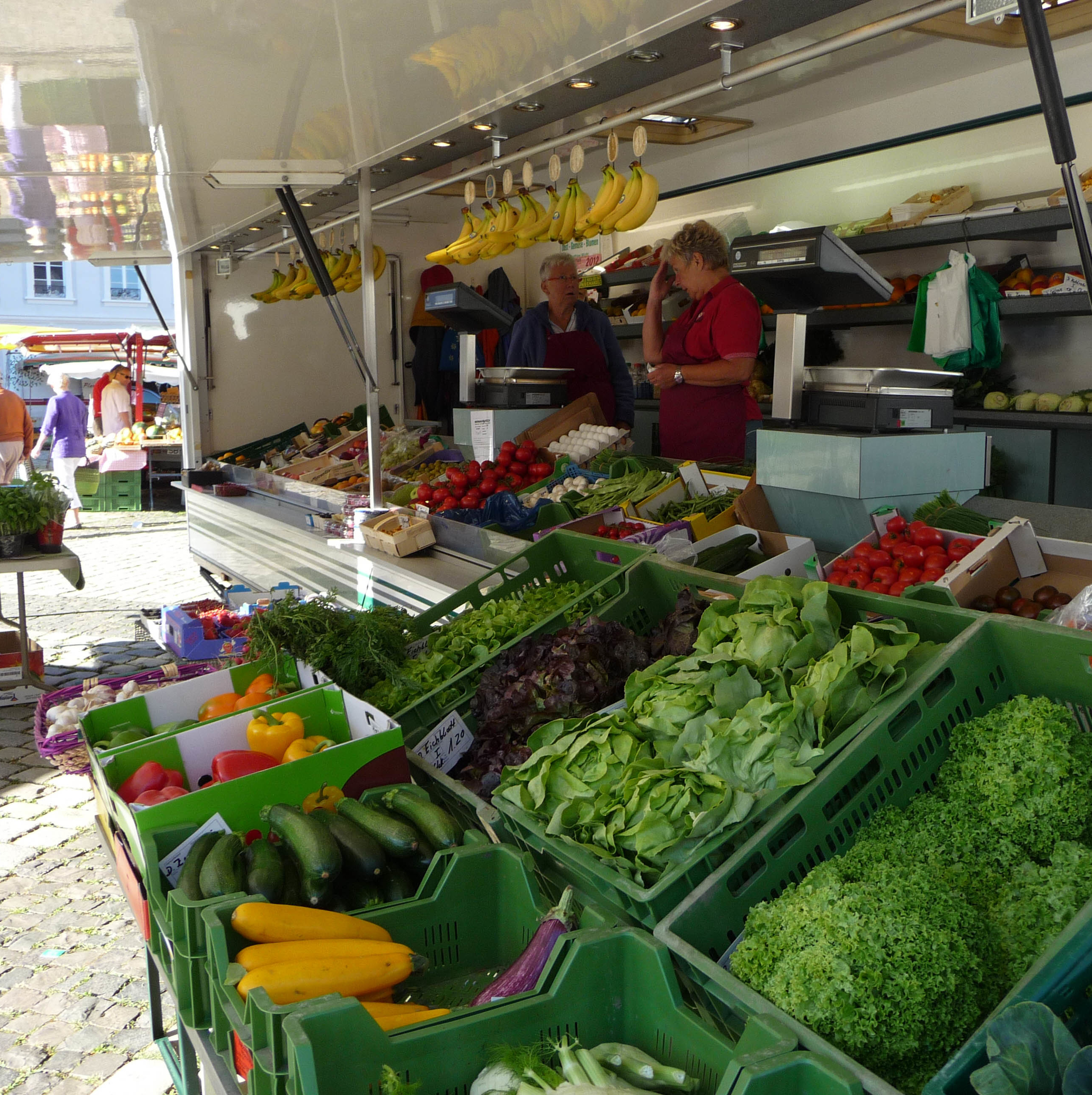 Gemüsestand mit Salatkisten und zwei Verkäufern. Im Hintergund der St. Johanner Markt in Saarbrücken.