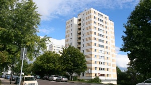 Hochhaus mit 12 Etagen an Straße, Fassade weiß und gelb. Bäume und Fahrzeuge.