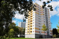 2 quadratische Hochhäuser mit 12 Etagen. Fassaden weiß. Farbakzente gelb/orange Haus vorne, hinten blau. Vorne hohe Bäume.