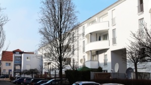 Hellgraue Gebäudezeile mit 4 Etagen, Balkone, im Winter. Vorne Parkplätze mit Fahrzeugen.