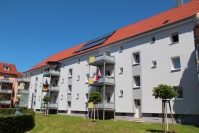 Hellgraues Wohnhaus mit neuem Dach, angebaute Balkone, 3 Etagen. Vorne Grünfläche und junge Bäume.