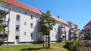 Hellgraues Wohnhaus mit neuem Dach, angebauten Balkone, 3 Etagen. Davor eine Wiese mit kleinen Bäumen.