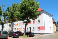 Hellgrauer Altbau mit drei Etagen, farbige Vierecke auf Fassade. Bäume und parkende Fahrzeuge. 