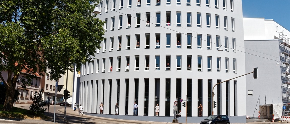 halbrundes Gebäude, 6 Etagen, hellgrau. Aus Fenstern schauen Menschen. Davor belebte Straße, links hoher Baum.