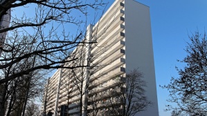 Hochhaus mit 15 Etagen, helle Fassade, Laubengänge. Haus ist 140 Meter lang. Vorne Gehweg, Straße, Bäume im Winter in der Morgensonne. 