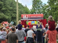 Eine Gruppe Menschen, darunter einige Kinder auf den Schultern von Männern sitzend, blickt auf eine Freilichtbühne auf der ein Theaterstück aufgeführt wird