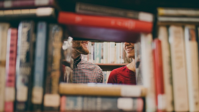 Zwei Personen lächeln sich durch eine Lücke zwischen den Büchern in einem Bibliotheksregal an.