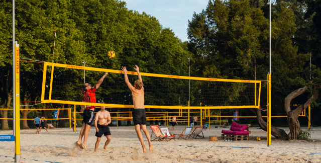 Drei Personen spielen Beachvolleyball im OneBeach. Im Vordergrund stehen zwei Liegestühle.