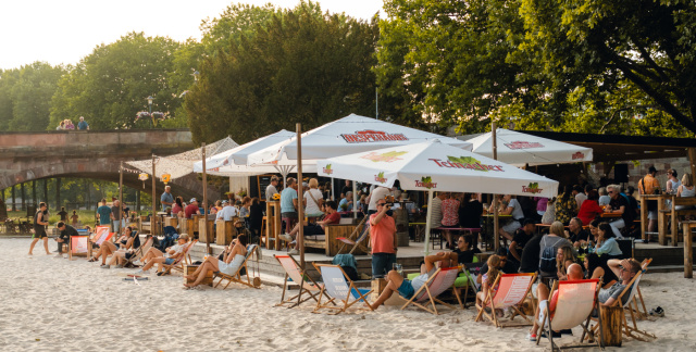 Im Vordergund ist ein künstlicher Sandstrand. In der Mitte sieht man Menschen in Liegestühlen unter Schirmen. Im Hintergrund links ist eine Brücke.