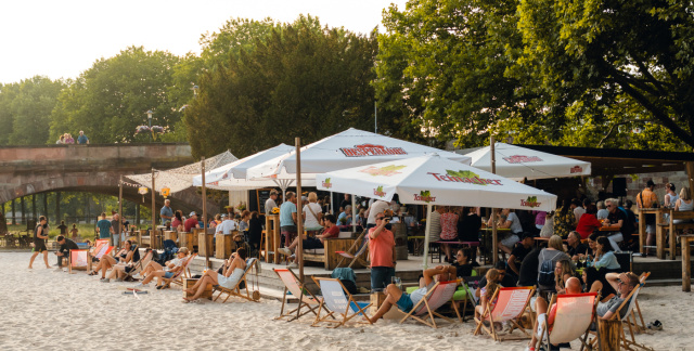 Im Vordergund ist ein künstlicher Sandstrand. In der Mitte sieht man Menschen in Liegestühlen unter Schirmen. Im Hintergrund links ist eine Brücke.