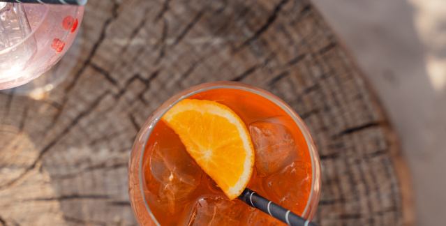 Von oben fotografiert: Ein Aperol Spritz steht mittig und ein Wildberry Lillet links oben auf einem Baumstumpf, welcher auf Sand steht.