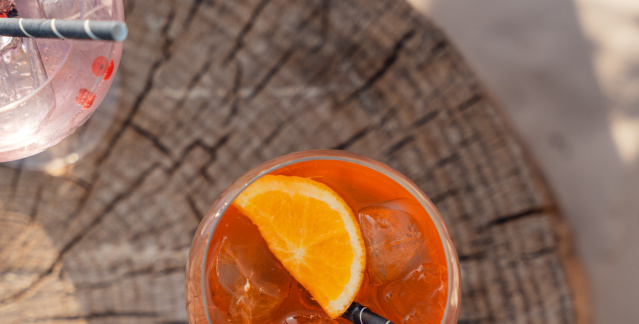 Von oben fotografiert: Ein Aperol Spritz steht mittig und ein Wildberry Lillet links oben auf einem Baumstumpf, welcher auf Sand steht.