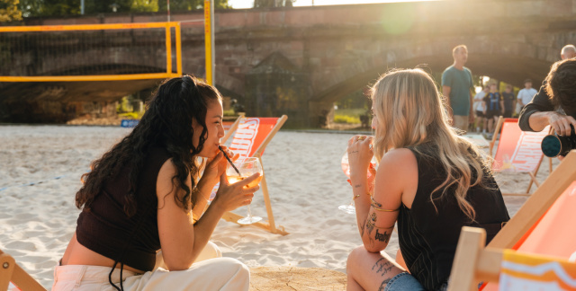 Im Vordergrund ist ein Teil von einem Liegestuhl. In der Bildmitte sitzen zwei Frauen in sommerlichen Outfits in Liegestühlen in der Sonne und trinken Getränke. Sie sind an einem künstlichen Strand. Im Hintergrund ist ein Beachvolleyballfeld und eine Brüc