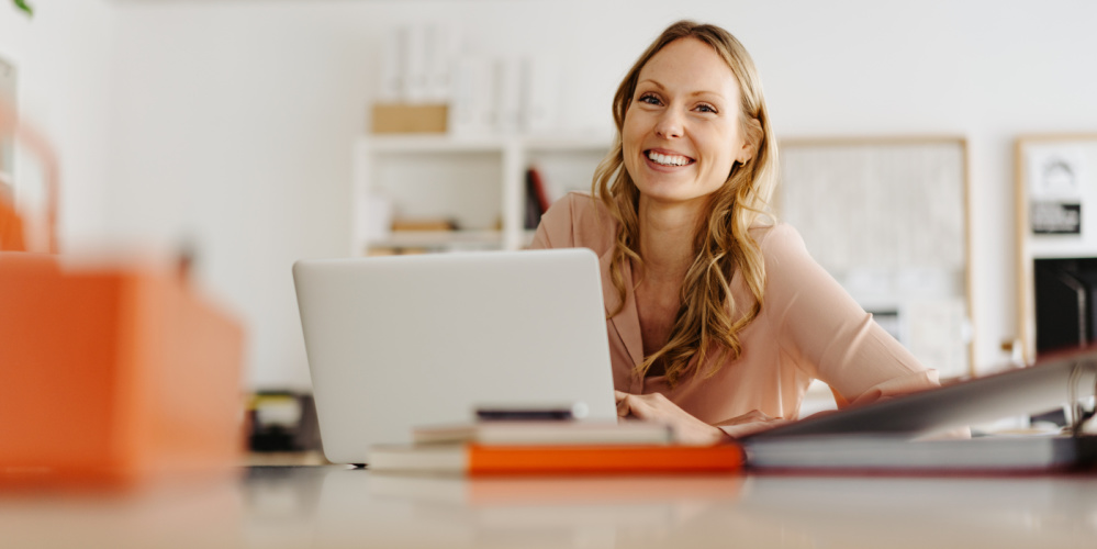 Eine lachende blonde Frau sitzt an einem Schreibtisch vor einem aufgeschlagenen Laptop. Foto: contrastwerkstatt / AdobeStock
