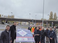 Vor dem Hintergrund einer Stadtautobahnbrücke liegt ein Schild um das sich einige Personen versammeln