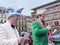 wei kostümierte Männer stehen auf einer Bühne, einer liest in ein Mikrofon, während der andere ein Blatt Papier in der Hand hält.