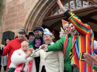 Ein Mann mit schwarzem Hut spricht in ein Mikrofon und blickt von der Bühne auf die versammelte Menge vor dem Rathausplatz.