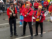 Drei Männer in roten Jacken und Narrenkappen stehen auf der Straße und halten große Konfettikanonen in den Händen.