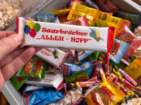 Eine Hand hält einen verpackten Schokoriegel mit der Aufschrift „Saarbrücker Alleh-Hopp“ über einer Kiste voller Süßigkeiten.