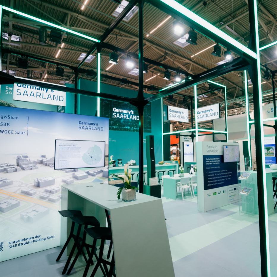 Ein offener mintgrün-leuchtender Messestand mit Hochtischen und -stühlen sowie Messewänden.